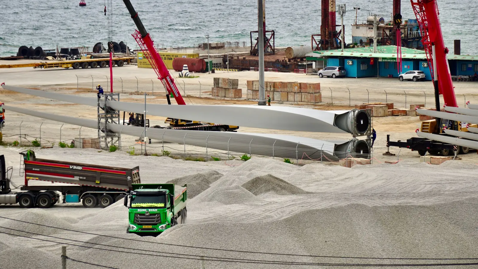 Palas de aerogenerador, grúas y camiones en una campa de obra junto al mar